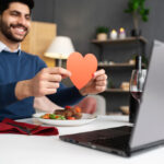 A man sitting at a table with a laptop, holding a paper heart and smiling, during an online date. The table is set with a meal, wine, and candles.