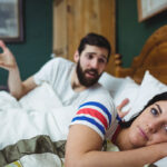 A couple in bed, with the woman lying on her side looking distant and the man sitting up, gesturing with concern and confusion.